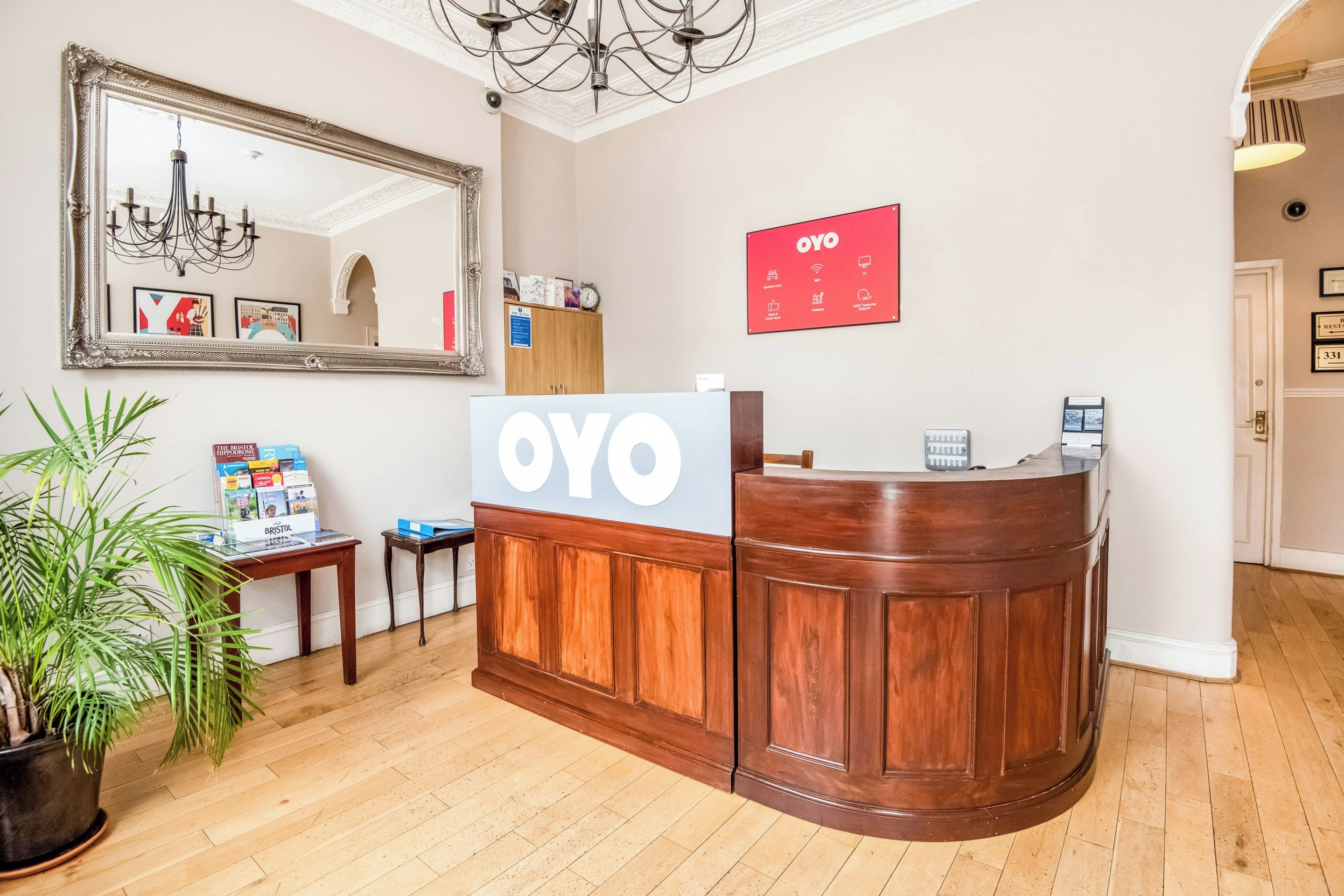 A reception desk with a sign reading 'OYO' and a large mirror above it. There is a potted plant to the left and a framed picture on the wall.