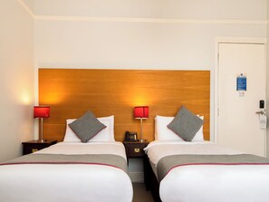 A hotel room with two beds, bedside tables, lamps, a door with a service sign, and a wall-mounted light fixture. - The Regency, Clifton Bristol (Bristol)