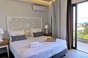A hotel room with a large bed, a decorative headboard, a nightstand with a lamp, and a view through a large window. - Villaggio Boutique Hotel Hersonissos - Adults Only (Hersonissos)