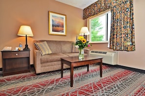 Ein Hotelzimmer mit einer Couch, zwei Lampen, einer Vase mit Blumen, einem gerahmten Bild, einem Fenster mit gemusterten Vorhängen und einem Teppich.