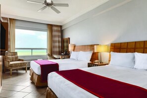 Quarto de hotel com duas camas, um ventilador de teto, vista para o exterior, uma escrivaninha com luminária e uma cadeira.