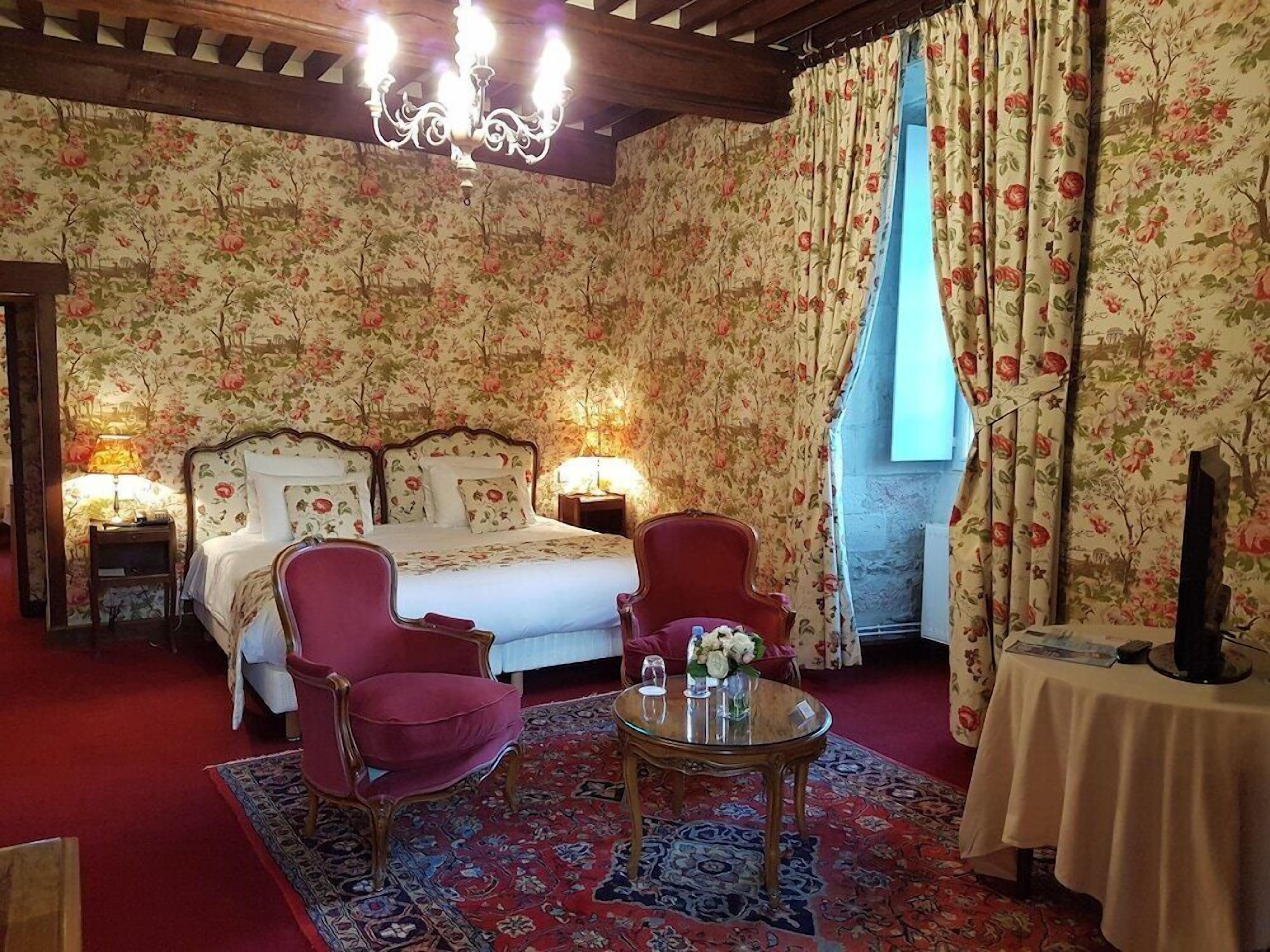 Habitación de hotel con cama doble, dos sillas, una mesa redonda, candelabro, papel tapiz floral y una ventana con cortinas.