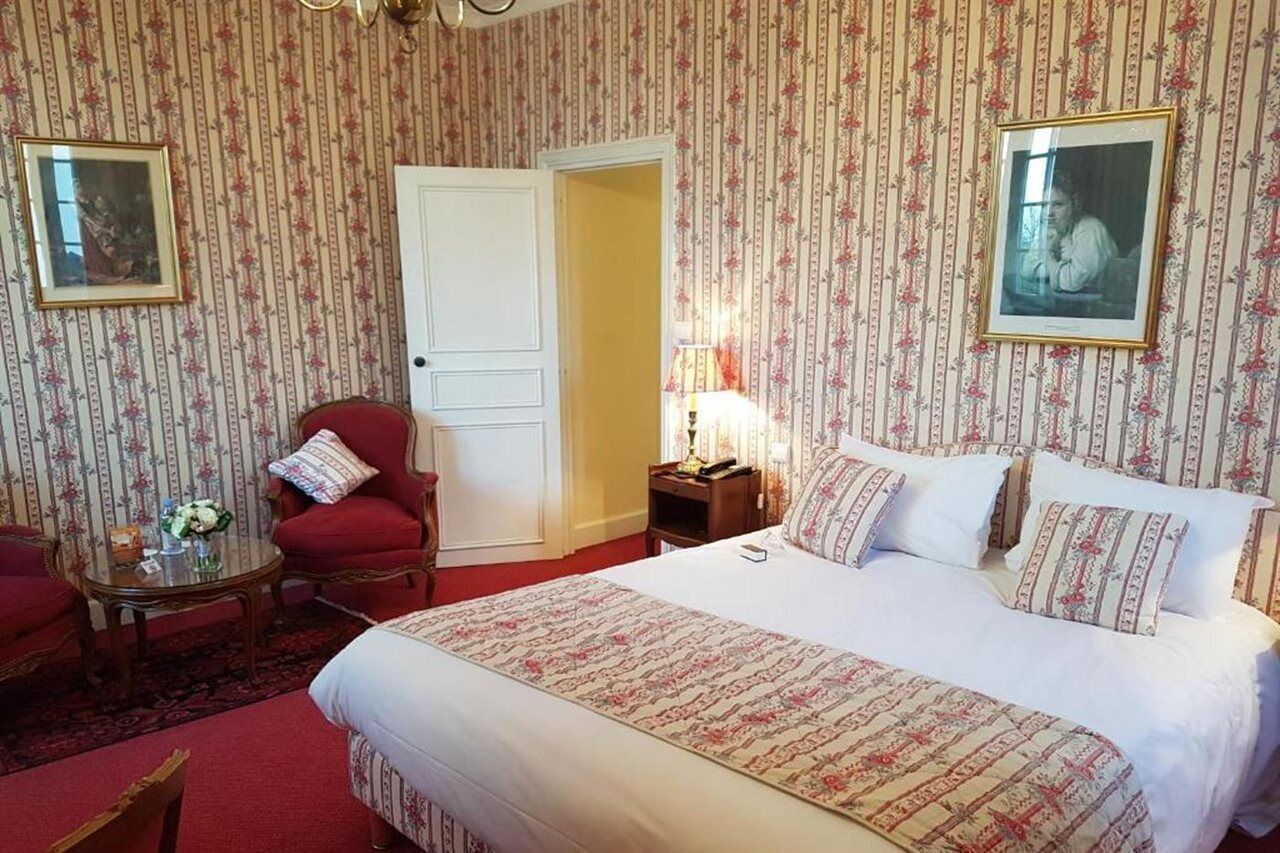Una camera d'albergo con un letto, una poltrona rossa, un tavolino con un vaso e una porta che conduce a un'altra stanza.