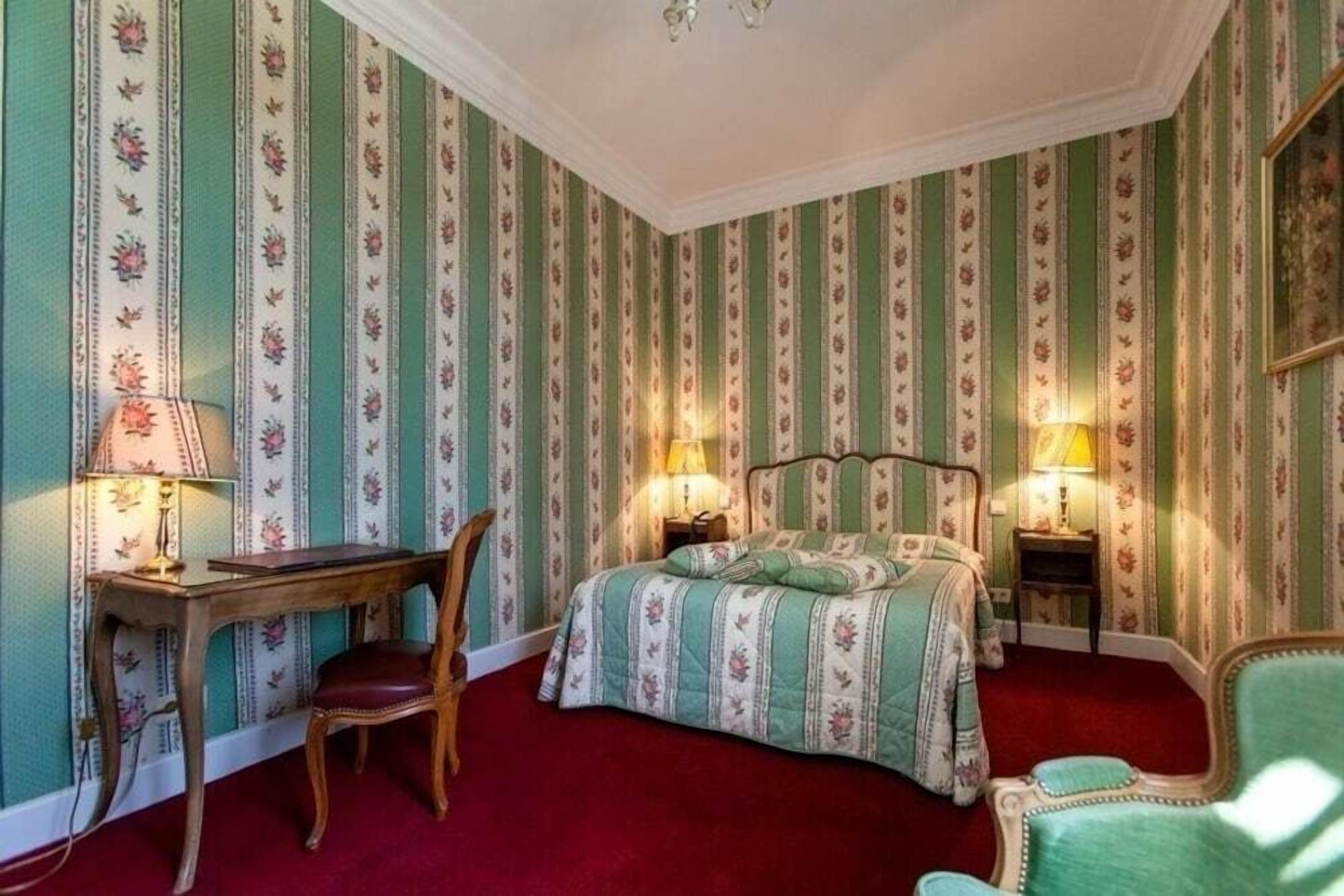 Ein Zimmer mit gestreifter Tapete, einem Bett mit Blumenmuster, zwei Nachttischlampen, einem Holztisch mit Stuhl und einem roten Teppich.