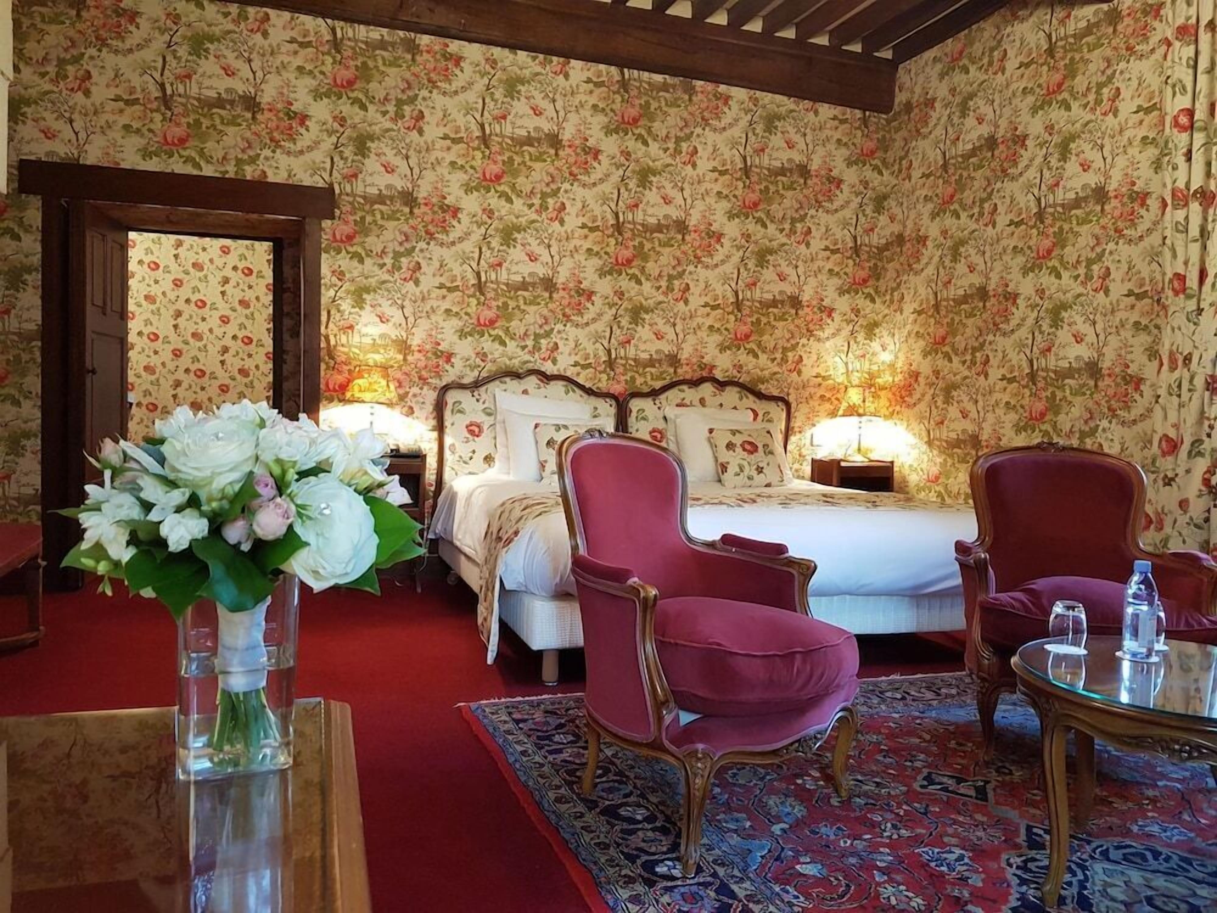 A hotel room with floral wallpaper, a bed with two pillows, a nightstand with a lamp, a red armchair, a round table with a glass top, and a vase with flowers.