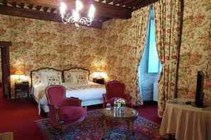 Ein Zimmer mit einem Bett, zwei Sessel, einem Tisch mit einer Vase und einer Kandelaber.