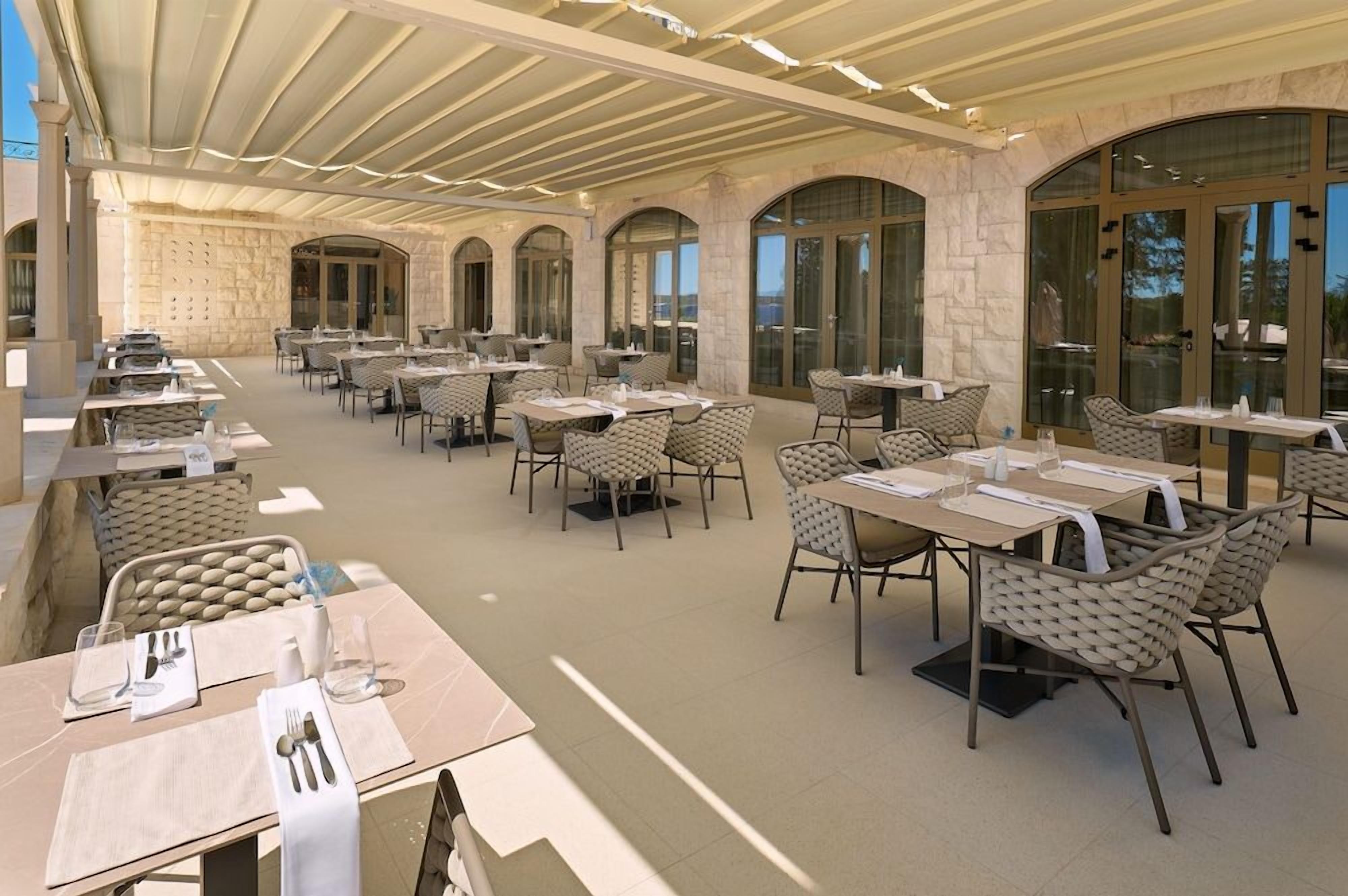 Un área de comedor al aire libre con varias mesas y sillas, paredes de piedra y ventanas grandes.
