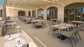 Un área de comedor al aire libre con varias mesas y sillas, paredes de piedra y ventanas grandes.