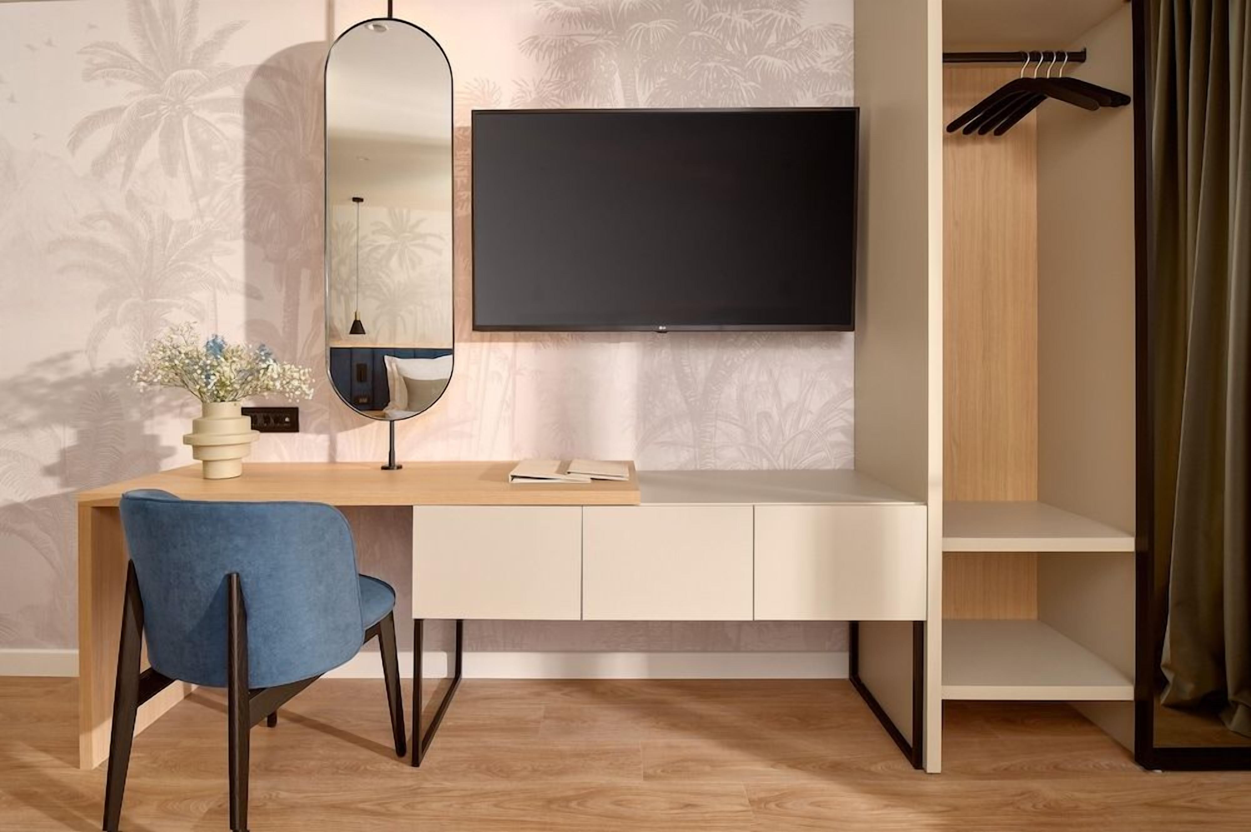 A modern room with a flat-screen TV mounted on the wall, a desk with a chair, a mirror, and a wardrobe with a hanging rack.