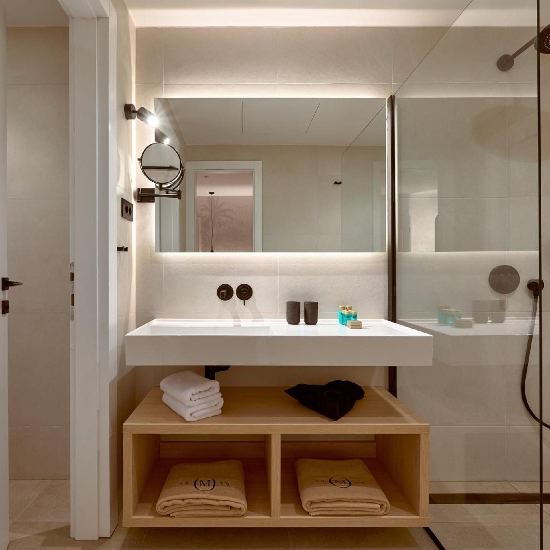 Un baño moderno con un lavamanos blanco, un espejo y una ducha. Hay toallas y artículos de aseo en el lavamanos.