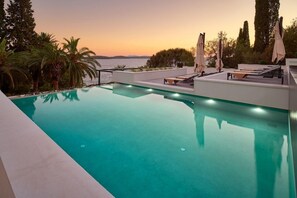 Une piscine dotée de chaises longues et de parasols, offrant une vue sur une étendue d’eau au coucher du soleil.