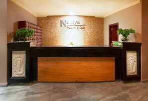 Vastaanottotiski, jossa on kyltti, jossa lukee ”Nadiya Health & Spa”. Tiskillä on molemmin puolin ruukkuistutettuja kasveja, ja lattia on marmoria.