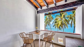 Un balcón con vistas a palmeras y al mar, una mesita pequeña, dos sillas de mimbre y una ventana con un mural tropical.
