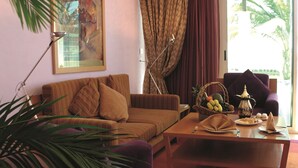 A living room with a sofa, a coffee table with fruit and a teapot, a painting on the wall, and a window with curtains. - Movenpick Resort Al Nawras Jeddah (Jeddah)
