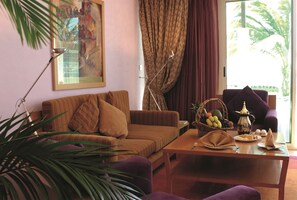 A living room with a sofa, a coffee table with fruit and a teapot, a painting on the wall, and a window with curtains. - Movenpick Resort Al Nawras Jeddah (Jeddah)