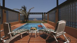 A covered outdoor swimming pool with two lounge chairs and a table, a palm tree, and a view of the sea. - Movenpick Resort Al Nawras Jeddah (Jeddah)