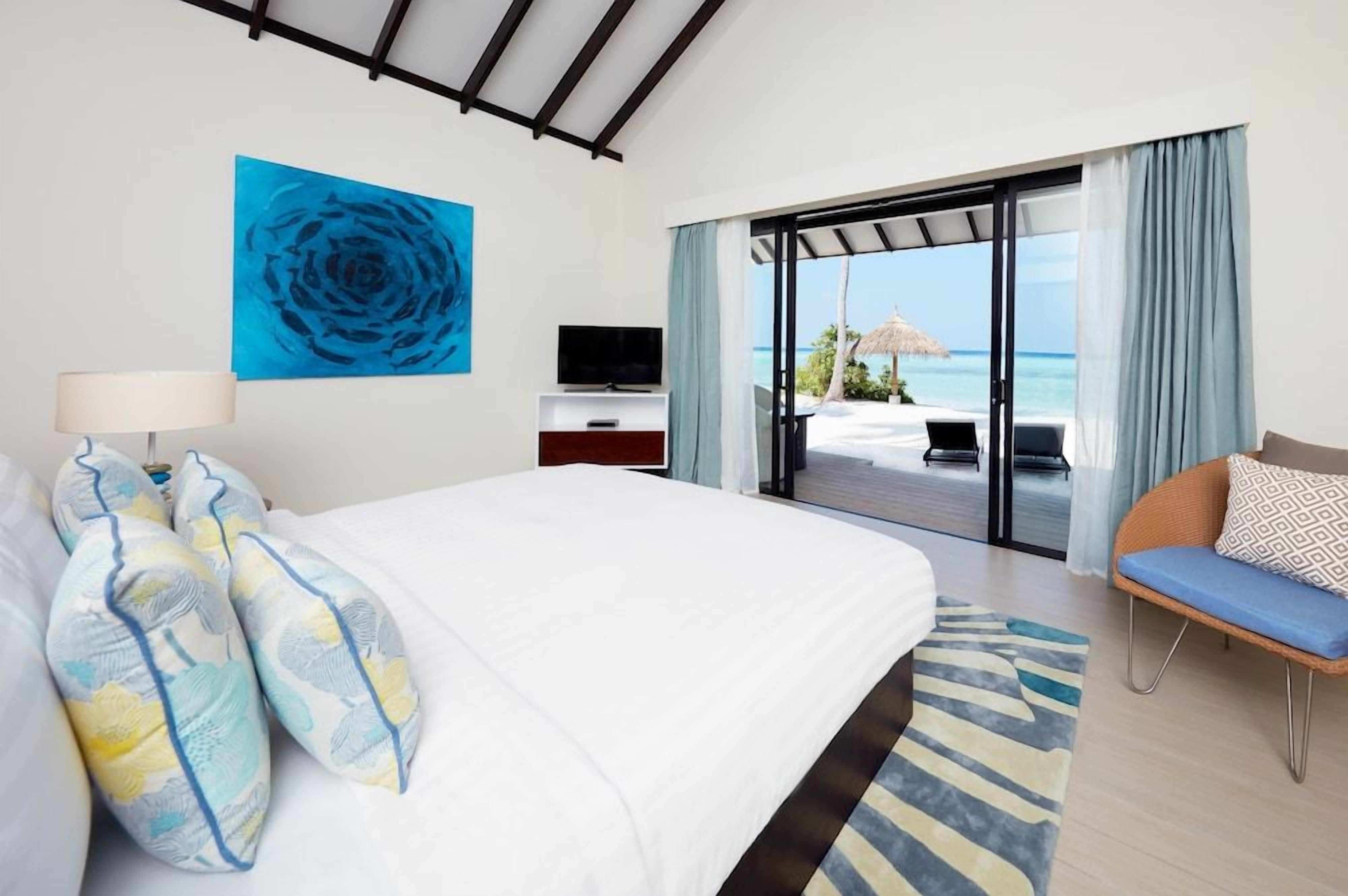 A hotel room with a bed, a TV, a view of the beach, and a balcony with a chair.