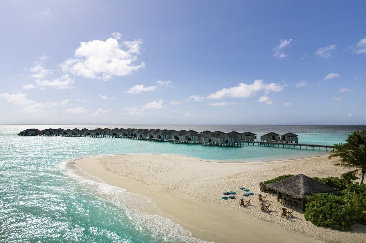 A beach resort with overwater bungalows connected by a pier, a sandy beach, and a clear blue sky.