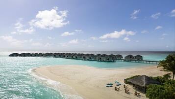 A beach resort with overwater bungalows connected by a pier, a sandy beach, and a clear blue sky.