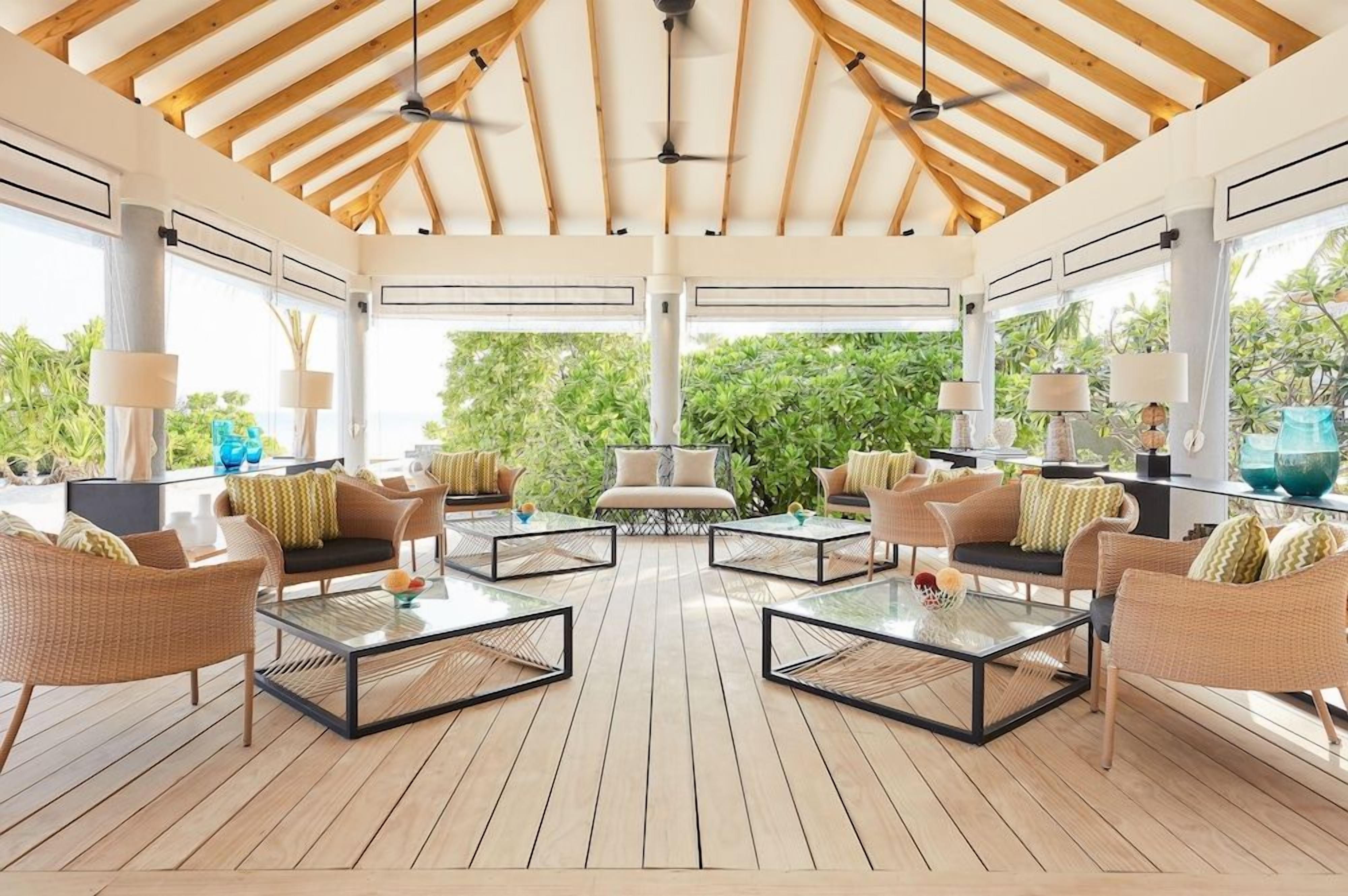 A covered outdoor seating area with wicker furniture, glass tables, and a view of greenery.