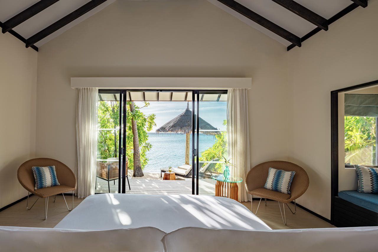 A bedroom with a large bed, two chairs, a glass table, and a view of the sea through a sliding glass door.