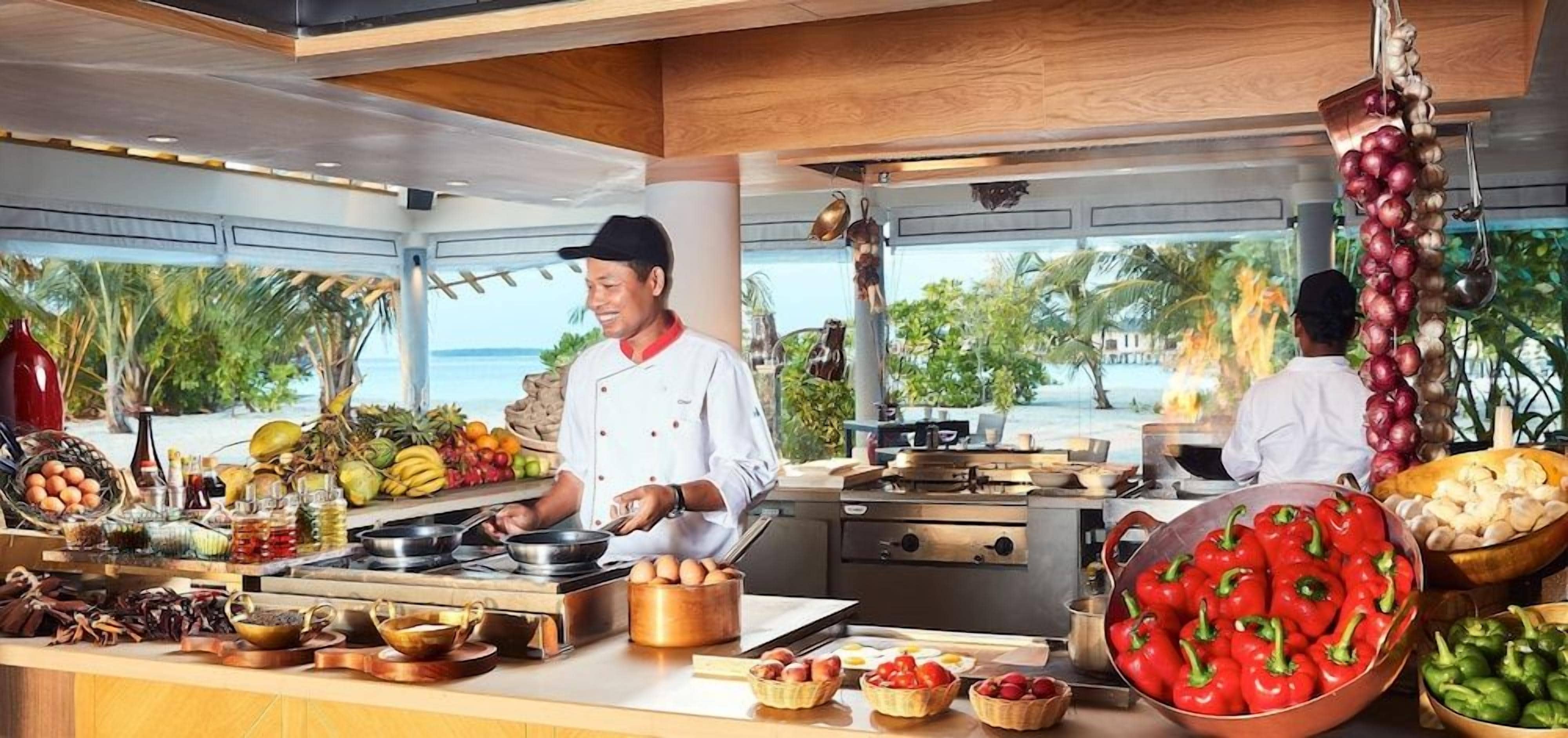 A kitchen with chefs preparing food, a view of the sea, and various fruits and vegetables on the counter.