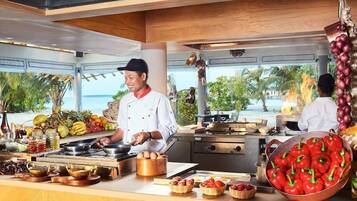 A kitchen with chefs preparing food, a view of the sea, and various fruits and vegetables on the counter.