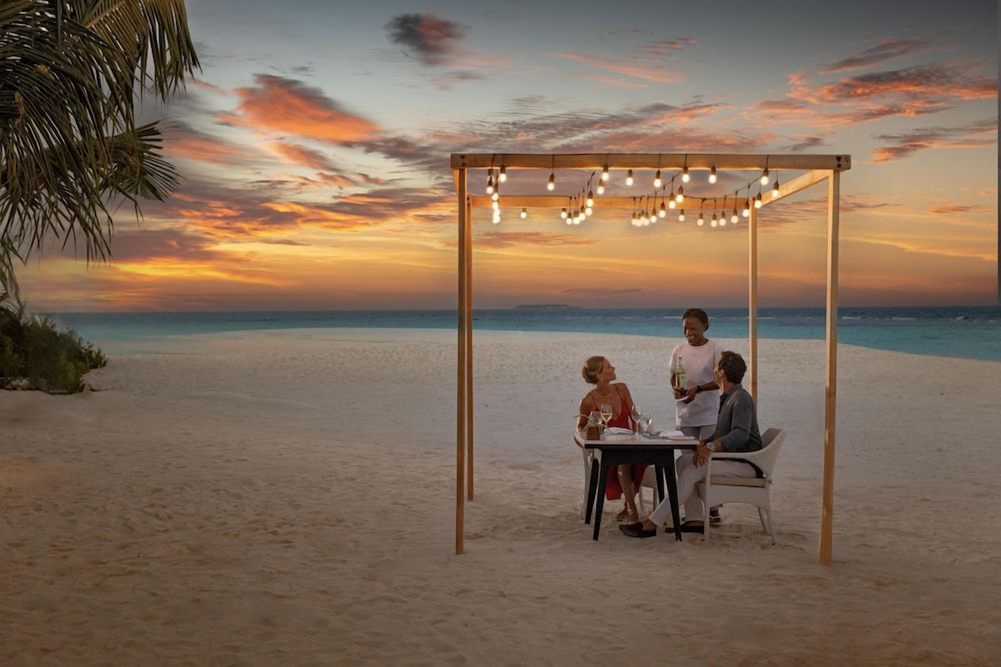 A beachside dining setup with a table and chairs under a pergola with string lights.