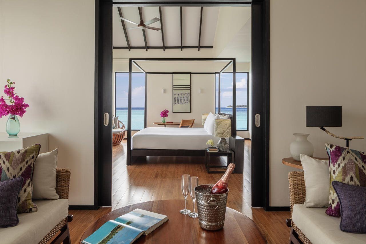 Habitación de hotel con cama, vista al mar, balcón y zona de comedor con mesa y sillas.