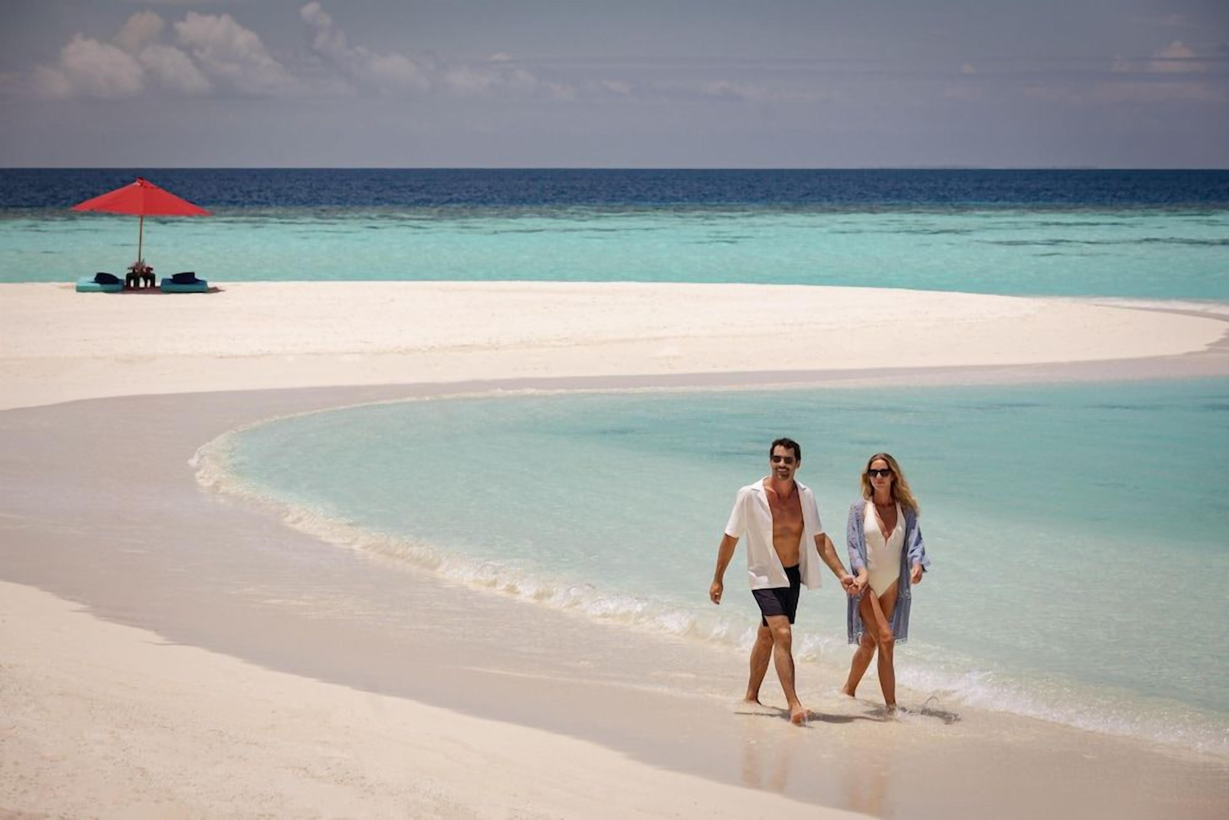 Un couple se promène sur une plage, avec un parasol rouge et des chaises longues en arrière-plan.