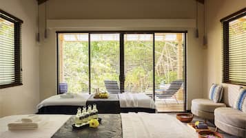 A spa room with two beds, a sofa, and a view of greenery through large windows.