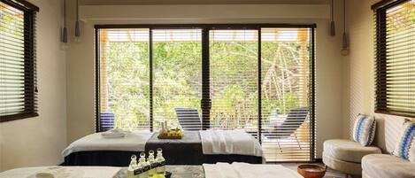 A spa room with two beds, a sofa, and a view of greenery through large windows.