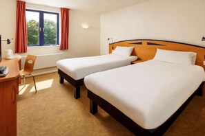 A hotel room with two single beds, a desk with a chair, a window with curtains, and a wall-mounted lamp. - Preston North (Preston)