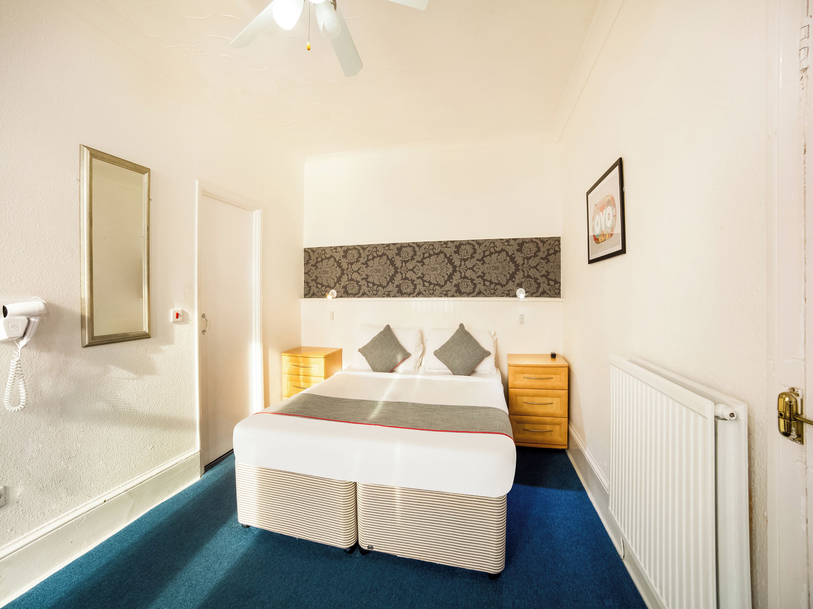 A hotel room with a bed, bedside tables, a mirror, a radiator, and a ceiling fan.