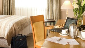 Una habitación de hotel con una cama, un escritorio con una computadora portátil, una lámpara, una cafetera y un florero con flores.