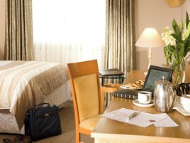 Una habitación de hotel con una cama, un escritorio con una computadora portátil, una lámpara, una cafetera y un florero con flores.
