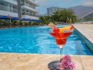 Ein Cocktail mit Obstbeilage auf einem Tisch am Pool, ein Schwimmbecken und ein Hotelgebäude im Hintergrund.