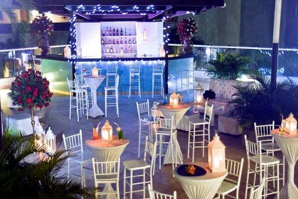 Uma área de bar ao ar livre com mesas, cadeiras, balcão bar com garrafas e iluminação decorativa.