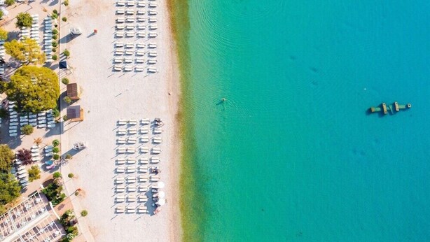 An aerial view of a beach with rows of sun loungers and umbrellas, a clear blue sea, and a small boat in the distance. - Maslinica Hedera Hotel (Labin)