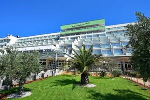 Um hotel moderno com fachada verde e branca, várias varandas e um jardim bem cuidado com palmeiras.