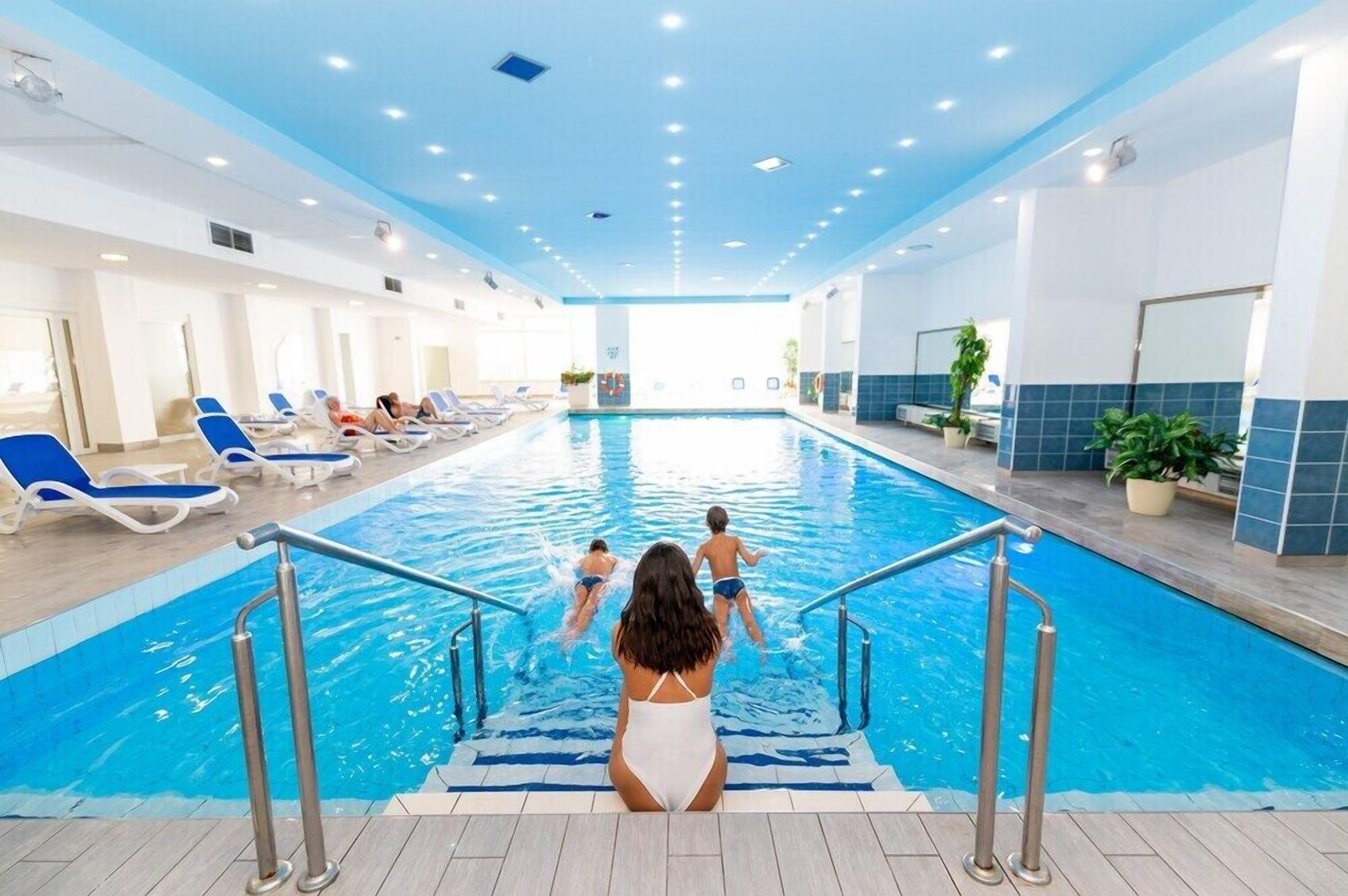 An indoor swimming pool with blue-tiled walls, a pool ladder, and lounge chairs along the sides.