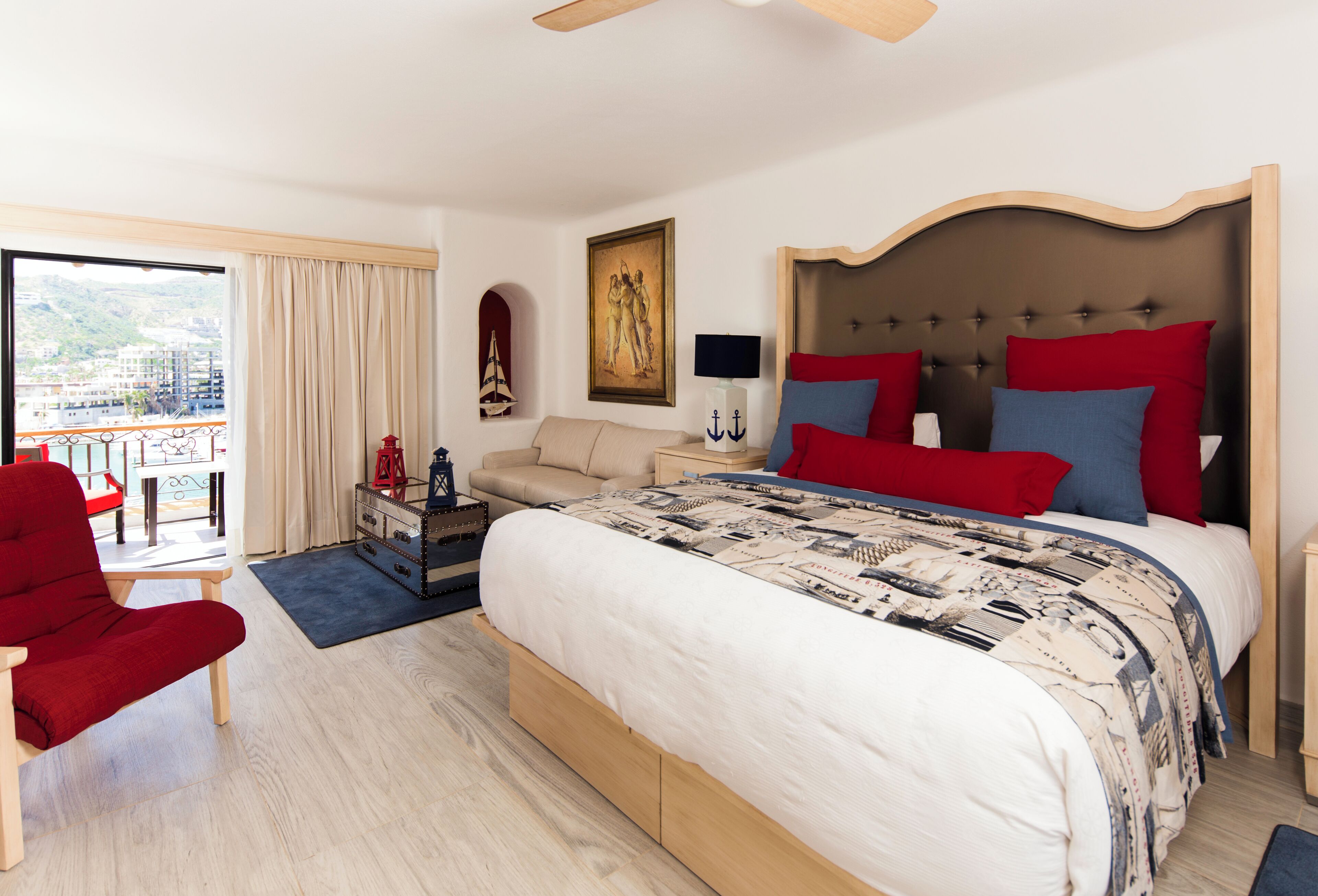 Habitación de hotel con una cama grande, un sillón rojo, una mesita, una mesita de noche, una lámpara, un cuadro y vistas a la ciudad.