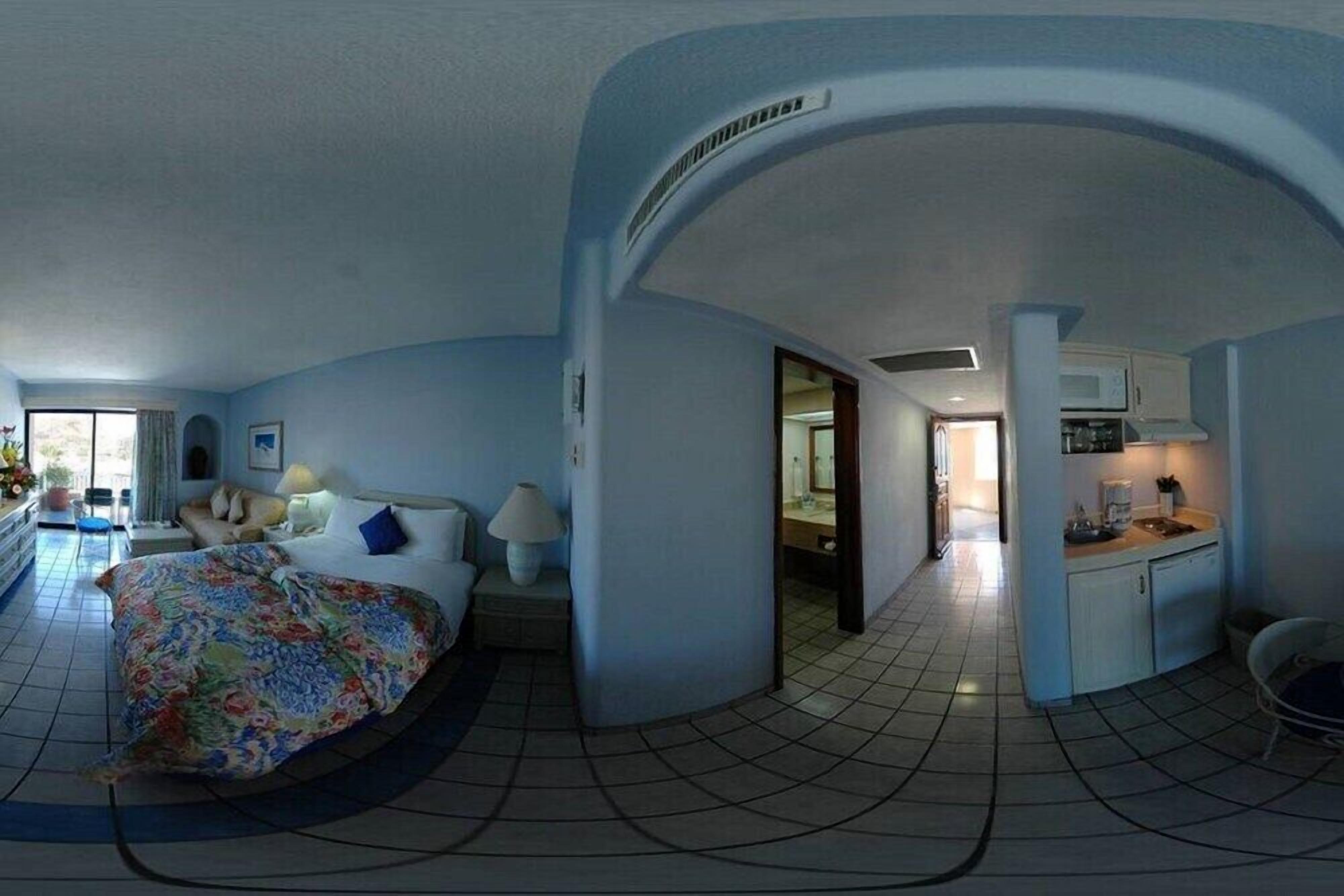 A hotel room with a curved ceiling, a bed with floral bedding, a bedside table with a lamp, and a view of the adjacent bathroom and kitchen area.