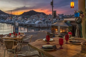 A waterfront restaurant with outdoor seating, a fireplace, and a view of boats and a mountain at sunset.
