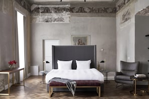 Quarto com cama grande, uma cadeira, mesa pequena, mesa de cabeceira e janela com cortinas.