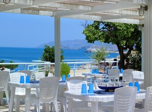 A seaside restaurant with white plastic chairs and tables, a view of the sea, and a boat docked nearby.