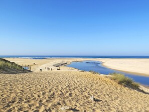A sandy beach with a clear blue sky, a small water body, and a few people walking. - Club Royal Océan La Prade by Interhome (Moliets)