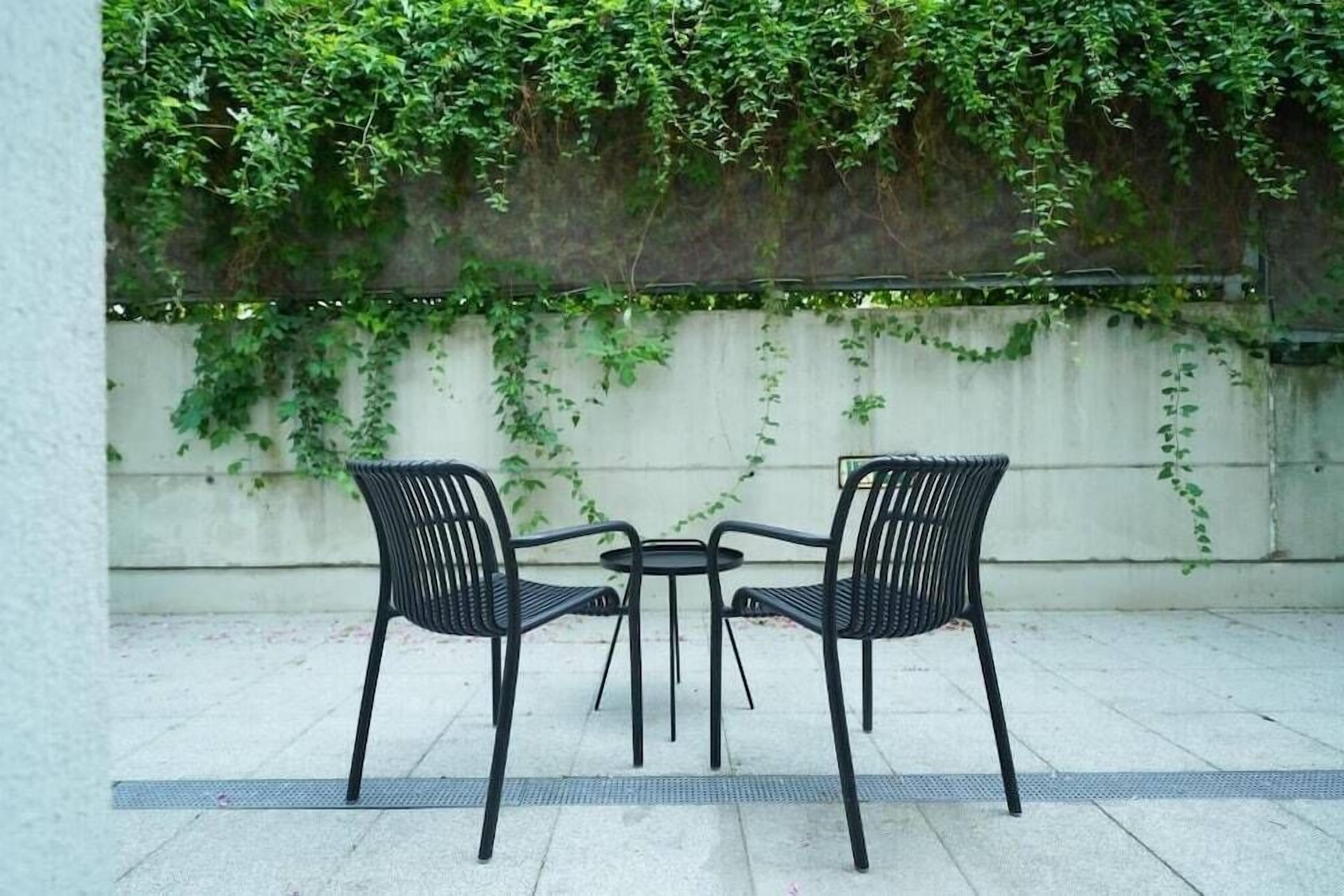 Zwei schwarze Metallstühle und ein kleiner runder Tisch auf einer gefliesten Terrasse mit weißer Wand und grünem Efeu im Hintergrund.