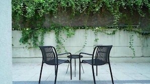Two black metal chairs and a small round table on a tiled patio with a white wall and green ivy in the background. - Smart Hotel Schonbrunn (Vienna)