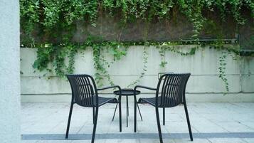 Zwei schwarze Metallstühle und ein kleiner runder Tisch auf einer gefliesten Terrasse mit weißer Wand und grünem Efeu im Hintergrund.
